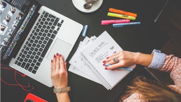 Frau sitzt vor einem Comöuter und hat eine Liste vor sich liegen. Eine Tasse Kaffee steht rechts von ihr. Rechts neben ihr liegen 4 Filzstifte in den Farben orange, gelb, rosa und blau.