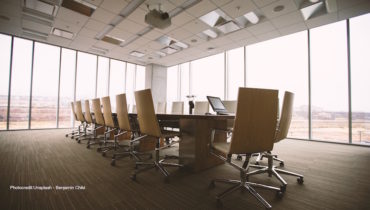 Ein Meetingtisch steht in der Mitte eines großen Büroraums. Darum herum sind weiße Bürostühle gestellt.