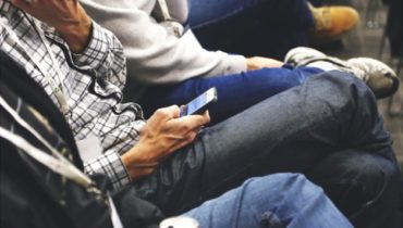 Mehrere Personen sitzen nebeneinander. Ein Mann hält ein Handy ein anderer eine Cola-Dose.