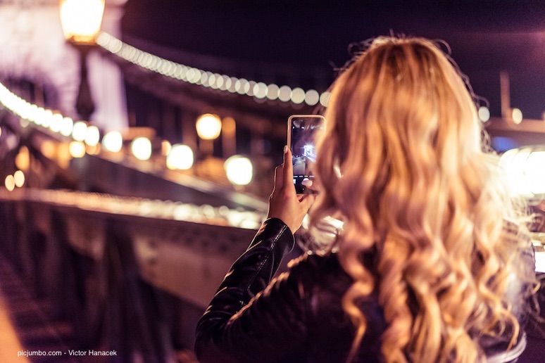Blondes Mädchen fotografiert in der Nacht mit ihrem Smartphone eine erleuchtete Brücke