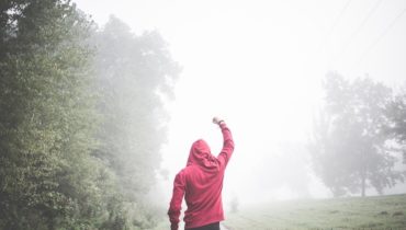 Ein Mann im roten Kaputzenpulli geht einen Weg entlang und streckt den Arm wie zum Sieg hoch. Links von ihm ist eine Baumreihe und rechts von ihm eine Wiese