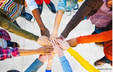 Arme sind in der Mitte des Bildes und die Hände legen sich übereinander. Die Arme haben verschiedene Hautfarben und beschreiben einen Zusammenhalt als Community.