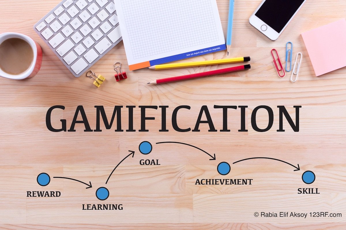 Holzplatte auf dem einen Tastatur liegt, ein Block, bute Stifte und ein Iohone. Darunter steht Gamification und Pfeile zeigen auf Reward, Learning, Goal, Achievement und Skill.